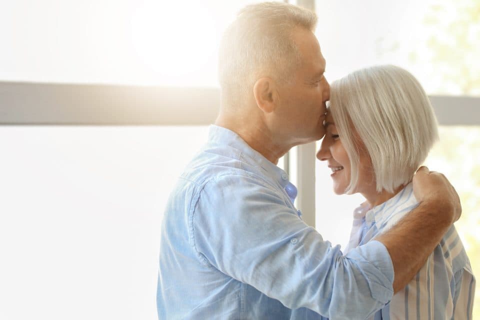 An older couple embracing, he kisses her forehead in soft daylight